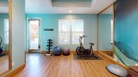 Located in La Quinta, California, this dedicated fitness studio is a thoughtfully designed workout space that combines functionality with stylish aesthetics. The room is finished with warm hardwood flooring throughout and painted in a calming aqua-blue palette that creates an energizing yet relaxed atmosphere. A tray ceiling with recessed lighting adds architectural depth and ensures the space is well-lit for exercise.

A striking floor-to-ceiling abstract mural in swirling shades of teal, white, and gold serves as a dramatic focal point on one wall, lending an artistic sensibility to the room. Large framed mirrors are strategically positioned along the walls to amplify the sense of space and support proper form during workouts. Natural light filters in through multiple windows fitted with blinds, offering a bright and inviting environment.

The studio is fully equipped with a wide array of fitness equipment, including an air resistance exercise bike, stability balls, a dumbbell rack, kettlebells, a medicine ball tower, resistance bars, yoga mats, and step platforms. Open shelving units provide organized storage for accessories and equipment. A wall-mounted television is also present, ideal for following workout classes or entertainment during training sessions. This space is a complete, private fitness retreat.