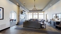 This single-floor space showcases a thoughtfully designed communal lounge area that blends elegance with comfort. The room's most striking feature is a floor-to-ceiling marble accent wall housing a sleek linear fireplace and a mounted flat-screen television, creating a bold and refined focal point. Flanking archways on either side of this feature wall add architectural character and provide graceful transitions to adjoining areas.

The soaring tray ceiling elevates the sense of grandeur, while a decorative beaded chandelier serves as a central design element, casting warm light throughout the room. Recessed lighting and track lighting complement the natural light that floods the space through a generous bank of tall windows along the far wall, offering a bright and airy atmosphere.

The seating area is anchored by a large, plush sectional sofa in a neutral gray tone, accompanied by additional upholstered seating and side tables arranged to encourage conversation and relaxation. The flooring transitions between large-format dark tile and area rugs, adding texture and visual depth. Overall, the space exudes a polished, contemporary aesthetic suited for sophisticated social gatherings.