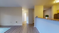 Located at 10906 E Portland Ave in Tacoma, Washington, this single-floor residence offers a well-organized layout with a clean, modern aesthetic. The living areas feature durable vinyl plank flooring in a warm wood tone, complemented by neutral gray and white walls that create a bright, open atmosphere. A bold yellow accent wall adds a touch of character near the kitchen pass-through, which features a granite-style countertop overhang ideal for casual dining.

The kitchen is efficiently designed in a galley style, outfitted with light maple cabinetry, granite-style countertops, tile backsplash, and stainless steel appliances including a refrigerator, electric range, and over-the-range microwave. Adjacent utility space provides additional storage and houses the water heater. The bedrooms are carpeted for comfort, with one room featuring a large picture window that frames views of mature evergreen trees and the surrounding neighborhood, along with a baseboard heater beneath. The bathroom is finished with matching light maple cabinetry, a granite-style vanity top, wood-look vinyl flooring, and a shower with curtain rod. Throughout, the space maintains a consistent, updated finish palette suited for comfortable everyday living.