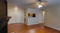 This single-floor space is designed with an open and functional layout that promotes a sense of connectivity between its living areas. The main living area features warm hardwood flooring that extends throughout, contributing to a cohesive and inviting atmosphere. A ceiling fan with an integrated light fixture is centrally positioned, providing both air circulation and ambient lighting.

A defining architectural feature is the pass-through window cut into the dividing wall between the living area and the kitchen, allowing visual openness and easy interaction between the two spaces. The kitchen is visible through this opening and is equipped with honey-toned wood cabinetry and black appliances, offering a practical cooking environment.

Adjacent to the living area, a set of double bi-fold closet doors provides generous storage capacity. The walls are finished in a neutral, light beige tone, complementing the warm flooring and creating a bright, welcoming interior. The textured ceiling adds a classic touch to the overall aesthetic.

A hallway visible beyond the central dividing wall suggests additional rooms, further enhancing the functional versatility of this well-organized, single-level space.
