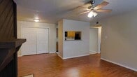 This single-floor space is designed with an open and functional layout that promotes a sense of connectivity between its living areas. The main living area features warm hardwood flooring that extends throughout, contributing to a cohesive and inviting atmosphere. A ceiling fan with an integrated light fixture is centrally positioned, providing both air circulation and ambient lighting.

A defining architectural feature is the pass-through window cut into the dividing wall between the living area and the kitchen, allowing visual openness and easy interaction between the two spaces. The kitchen is visible through this opening and is equipped with honey-toned wood cabinetry and black appliances, offering a practical cooking environment.

Adjacent to the living area, a set of double bi-fold closet doors provides generous storage capacity. The walls are finished in a neutral, light beige tone, complementing the warm flooring and creating a bright, welcoming interior. The textured ceiling adds a classic touch to the overall aesthetic.

A hallway visible beyond the central dividing wall suggests additional rooms, further enhancing the functional versatility of this well-organized, single-level space.