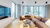 This expansive, single-level amenity space is thoughtfully designed to accommodate both social gatherings and relaxed lounging. The open-plan layout is divided into two distinct functional zones: a dining area and a lounge area, connected seamlessly by wide-plank light hardwood flooring that runs throughout the entire space.

The dining zone features a large, dark-topped communal table surrounded by a mix of upholstered and wooden chairs, complemented by a sleek bar counter with integrated cabinetry and pendant lighting overhead. Decorative vases with dried botanicals add warmth to the contemporary aesthetic.

The lounge zone is anchored by a generously sized sectional sofa paired with a low coffee table, positioned in front of a wall-mounted flat-screen television. A distinctive exposed concrete cylindrical column serves as an architectural focal point, adding a refined industrial character to the otherwise clean, white-walled interior.

Floor-to-ceiling curtain-wall glazing wraps the exterior perimeter, flooding the space with abundant natural light and framing sweeping urban skyline views. Full-height glass doors provide direct access to an outdoor terrace. Cylindrical black pendant fixtures mounted to the white ceiling provide consistent, layered artificial lighting throughout.