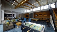 This vibrant recreation lounge is designed for socializing and entertainment, combining contemporary design with a welcoming, energetic atmosphere. The space immediately impresses with its soaring ceilings adorned with exposed wood beams and abundant clerestory windows that line the upper walls, flooding the interior with natural light while adding architectural character.

At the heart of the space, a foosball table and a pool table serve as focal points for recreation, complemented by stylish blue accent chairs and a comfortable lounge seating area with a modular sofa and colorful throw pillows. A low-profile cube ottoman anchors the lounge zone, creating an inviting setting for casual conversation or relaxation.

Toward the back, a dining area with chairs and a large wall-mounted flat-screen television provides a comfortable setting for group gatherings or watching events. Additional mounted screens are visible throughout, ensuring entertainment is accessible from multiple vantage points. The warm amber-toned accent wall adds depth and contrast to the space, while the combination of recessed and pendant lighting enhances the ambiance during evening hours.

The overall design balances an industrial-modern aesthetic with warm, residential touches, making this a dynamic communal space suited for both lively recreation and relaxed socializing.