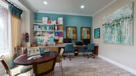 The Business Center is a thoughtfully designed, single-floor space that combines functionality with a lively aesthetic. Accessed through a set of glass-paneled French doors, the center is visually open and connects seamlessly to the broader common area beyond, which features soaring ceilings, light wood-look flooring, and abundant natural light streaming through large windows with teal and cream accent drapery.

Inside the business center, a bold teal accent wall sets an energetic tone, contrasted by neutral walls and crown molding throughout. The workstation area along the back wall offers multiple computer terminals and a printer, with teal ergonomic chairs providing comfortable seating. A built-in pegboard and open shelving system displays an extensive array of office and craft supplies, keeping resources organized and accessible.

A round wooden table with upholstered seating provides a collaborative space for informal meetings or individual use. Complimentary Wi-Fi is prominently advertised. Framed artwork and abstract paintings adorn the walls, enhancing the creative atmosphere. Light wood-look flooring ties the space together, contributing to a clean, modern, and welcoming environment suited for productivity.