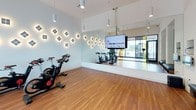 This contemporary fitness facility is thoughtfully designed to support cycling-focused workouts in a sleek, well-appointed environment. The space is anchored by a dedicated spin studio housing multiple Peloton stationary bikes arranged for group or individual sessions. The studio features warm hardwood flooring throughout, creating a cohesive and inviting aesthetic.

A striking design element is the array of diamond-shaped decorative wall sconces installed across the studio walls, each emitting a warm glow that adds both ambiance and visual interest. Full-length mirrors line portions of the walls, enhancing the sense of space and allowing users to monitor their form during workouts. A wall-mounted flat-screen television is positioned within the studio, providing access to on-demand fitness content.

Floor-to-ceiling glass walls with bold yellow and amber accent panels define the perimeter of the spin area, offering views to an outdoor terrace and allowing natural light to fill the space. Pendant and recessed lighting supplement the natural light, maintaining consistent brightness throughout. The overall atmosphere is modern and energizing, combining functional fitness amenities with polished architectural details.