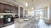 Located in Waterside Village, Houston, TX, this one-bedroom, two-bathroom residence offers a thoughtfully designed single-level layout with modern finishes and efficient use of space. The open-concept kitchen and living area serves as the heart of the home, featuring rich dark cabinetry, granite countertops, a breakfast bar island with pendant lighting, and black appliances including a full-size refrigerator, range, and microwave. Light wood-style flooring runs throughout, complementing the neutral color palette. The living area is bright and airy, benefiting from multiple windows and a sliding glass door that opens to a private balcony, bringing in natural light and outdoor views. A ceiling fan adds comfort to both the living area and bedroom. The primary bedroom is comfortably sized with a large window, ample closet space, and direct access to the en-suite bathroom. The en-suite bath features a dark wood vanity with granite countertops and a soaking tub with tile surround. A second full bathroom provides added convenience. Additional highlights include in-unit laundry and multiple closets for generous storage. The overall ambiance is clean, modern, and functional.