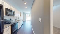 This single-floor unit presents a clean, modern design highlighted by consistent grey-toned hardwood-style flooring that runs throughout the main living areas, creating visual continuity and a cohesive aesthetic. The kitchen is designed with efficiency and style in mind, featuring white cabinetry with open shelving, granite countertops, a stainless steel sink with a gold-toned faucet, and black stainless appliances including a refrigerator, dishwasher, and range. Recessed lighting illuminates the kitchen corridor, which flows naturally into the open living area. The living space is bright and airy, benefiting from a large sliding glass door that connects the interior to an outdoor balcony, allowing abundant natural light and views of surrounding greenery. A second room, accessible via its own sliding glass door to the balcony, offers a versatile layout with clean white walls and overhead storage. The bathroom maintains the same refined palette, featuring large-format tile flooring, a granite-topped vanity with white cabinetry, a well-lit mirror with wall sconces, and a combined shower-tub area. Throughout, the design emphasizes simplicity, functionality, and modern detailing.