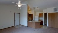This single-floor space offers an open-concept layout that seamlessly connects the living area to the kitchen, creating a sense of spaciousness and ease of movement. The living area is highlighted by dramatic vaulted ceilings, a ceiling fan with light fixture, and neutral carpeting that adds warmth to the space. Natural light fills the room, enhancing the airy feel.

The kitchen is well-appointed with warm wood-toned cabinetry, a white refrigerator, and a curved peninsula breakfast bar that provides additional counter space and serves as a natural divider between the kitchen and living areas. A double sink is integrated into the peninsula countertop, adding functionality to the layout. Track lighting above the kitchen ensures ample illumination for meal preparation. An in-unit washer and dryer area is visible, offering added convenience. A doorway leads to an additional room beyond the kitchen, suggesting further private living space. The entry door is positioned adjacent to the living area, providing a straightforward and welcoming entry point into the home. Overall, the space combines practical features with an open, comfortable design aesthetic.