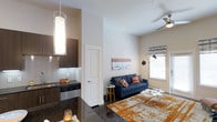 This thoughtfully designed single-floor space showcases a seamless open-concept layout that connects the kitchen and living area, creating an inviting and functional environment. The kitchen features dark espresso cabinetry, sleek granite countertops, stainless steel appliances, and a subway tile backsplash, all illuminated by a striking cylindrical pendant light that adds a modern touch. The living area is warm and welcoming, anchored by a bold navy blue sofa accented with colorful throw pillows and complemented by a vibrant orange area rug that adds depth and character to the space. A glass-top coffee table and a contemporary arc floor lamp contribute to the room's modern aesthetic. Natural light filters in through a window dressed with roman shades, brightening the crisp white walls adorned with tasteful wall art. The soaring ceilings enhance the sense of openness, while a ceiling fan ensures year-round comfort. Dark polished flooring runs throughout, tying the kitchen and living spaces together cohesively. The overall ambiance is modern yet comfortable, blending bold design choices with practical living elements to create a stylish and functional home environment.