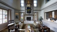 This refined single-floor space makes an immediate impression with its dramatic double-height ceilings that create an expansive, airy atmosphere. The living area is anchored by a grand white brick fireplace that rises toward the ceiling, framed by detailed millwork and topped with a large-scale landscape painting that draws the eye upward. Tall, slender windows flank the fireplace on both sides, flooding the room with natural light while offering views of the surrounding greenery. Leaded glass accent windows add a touch of classic architectural character to the walls. The interior design palette is composed of warm neutrals — soft grays, taupes, and earthy greens — carried through the walls, drapery, and furnishings. Symmetrically arranged sofas and upholstered seating are positioned around a rustic wood coffee table atop a richly patterned area rug, creating a welcoming and balanced gathering space. Side tables with decorative accents and potted greenery add warmth and life to the room. The overall ambiance is one of timeless elegance, combining traditional architectural details with thoughtfully curated furnishings to deliver a space that feels both grand and inviting.