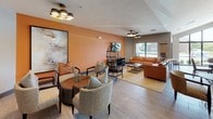 This single-floor community clubhouse is thoughtfully designed to offer a welcoming, multifunctional gathering space. The layout seamlessly connects several distinct activity zones, each with its own character while maintaining a cohesive modern aesthetic throughout.

The game area features a full-size billiard table set beneath striking circular pendant lighting, with large floor-to-ceiling windows that flood the space with natural light and provide open views of the surrounding greenery and parking area. Nearby, a round dining table with upholstered leather chairs creates an intimate spot for casual conversation or games.

A warm and inviting lounge area is anchored by bold burnt-orange accent walls, a wall-mounted flat-screen television, and rich cognac leather sofas arranged around a metal-framed coffee table atop a patterned area rug. Stylish cluster pendant chandeliers and a distinctive cage-style accent chair add personality and depth to the space.

The café-style bar station along the back wall features wood-paneled cabinetry, beverage equipment, and a digital display screen, serving as a central hub for refreshments. Ample seating is distributed throughout, with a variety of table and chair configurations suited for both solo and group use.

Light wood-toned tile flooring runs consistently throughout, unifying the open-plan layout. Together, the thoughtful furnishings, layered lighting, and vibrant color palette create a lively yet comfortable environment ideal for community gatherings and everyday relaxation.