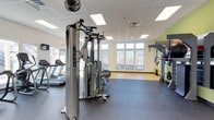 This fitness center is designed to support a comprehensive workout experience within a clean, functional layout. The space features durable rubber flooring throughout, providing a safe and resilient surface ideal for a range of exercise activities. A multi-station cable machine serves as a centerpiece for strength training, complemented by additional resistance equipment and TRX suspension training systems mounted along the wall. Cardio equipment, including treadmills, lines one side of the room, offering options for both warm-up and sustained aerobic workouts. Large windows along the far wall flood the space with natural light and offer views of the surrounding neighborhood, creating an open and energizing atmosphere. The drop-panel ceiling is fitted with recessed fluorescent lighting, ensuring bright and even illumination throughout. A ceiling-mounted fan provides additional airflow for comfort during intense sessions. The walls feature a neutral palette with subtle accent colors, contributing to a motivating yet calming environment. Overall, this fitness center is thoughtfully arranged to accommodate multiple users simultaneously while maintaining a spacious and well-organized feel.