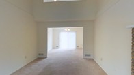 This two-floor space is defined by its impressive vaulted ceilings, which lend a sense of grandeur and openness to the main living area. The expansive vertical height draws the eye upward, enhancing the overall sense of scale. Neutral-toned walls and wall-to-wall carpeting in a warm beige hue run throughout, creating a cohesive and understated aesthetic that provides a versatile backdrop for furnishings.

The main living area flows seamlessly into an adjoining room through a wide open archway, offering an open-plan layout that encourages connectivity between spaces. The adjoining room features vertical blinds covering a large window, allowing diffused natural light to fill the interior with a soft, bright ambiance. A modest chandelier is visible through the archway, adding a subtle decorative element to the secondary space.

Flush-mounted HVAC vents are positioned along the baseboards in multiple locations, ensuring efficient climate control throughout. The clean, uncluttered walls and simple architectural lines give the space a calm, neutral character, making it highly adaptable to a variety of interior design styles.
