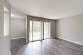 This single-floor, 1-bedroom, 1-bathroom residence offers a thoughtfully organized layout that maximizes its compact footprint. The living room is a standout space, featuring wood-style plank flooring, neutral gray walls, and a sliding glass door framed by sheer vertical blinds that floods the room with natural light and provides direct access to an outdoor area with greenery views. Adjacent to the living area, the galley-style kitchen is efficiently designed with white cabinetry, granite-look countertops, a subway tile backsplash, a stainless steel sink, and black appliances including a refrigerator and range, all set on light hardwood-style flooring. Two hallways connect the living spaces to the private areas of the home, with wall-mounted lighting providing a clean, functional aesthetic. The bedroom is a generously sized retreat carpeted in neutral tones, offering a window with exterior views and ample wall space. The bathroom features a full-size soaking tub with white subway tile surround, wood-style flooring, and a clean, minimal design. Throughout the unit, consistent neutral wall tones, white trim, and cohesive flooring choices create a unified, move-in-ready atmosphere.