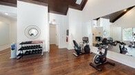 Located at 267–269 Charlotte Hwy in Asheville, NC, this well-appointed fitness center offers a bright, inviting environment for exercise and wellness. The single-floor space is defined by its striking vaulted ceilings with rich, dark wood detailing that draw the eye upward and create an airy, open atmosphere throughout.

Rich hardwood flooring runs the length of the space, lending warmth and a polished aesthetic. Large windows line multiple walls, flooding the interior with natural light while offering views of the lush outdoor surroundings, including a nearby pool area visible from the cardio section. The generous ceiling height enhances the sense of spaciousness and comfort during workouts.

The fitness center is equipped with a comprehensive selection of cardio machines, including multiple treadmills, ellipticals, and both upright and recumbent stationary bikes. A dedicated free weight area features a full rack of dumbbells, catering to strength training needs. Full-length mirrors are strategically placed to expand the visual space and assist with form during exercise.

Glass doors provide direct outdoor access, connecting the interior seamlessly to the exterior grounds. The clean, white walls and well-maintained equipment contribute to a professional and welcoming fitness environment, making this a functional and attractive amenity for the Asheville community it serves.