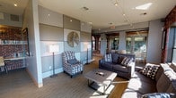 The Upper Clubhouse is a thoughtfully designed amenity space that seamlessly blends industrial character with contemporary comfort. Exposed red brick walls serve as a defining architectural element throughout, providing a warm, textured contrast to the sleek gray paneled accent walls adorned with modern metallic wall art and track lighting overhead.

The main lounge area features generous seating with plush gray sofas and a patterned accent chair arranged around a low-profile coffee table, creating an inviting space for relaxation or casual conversation. Large, multi-pane windows line the brick walls, flooding the interior with abundant natural light and offering open views of the surrounding treetops and skyline.

A dedicated media zone includes a wall-mounted flat-screen television and coordinating media console, ideal for communal viewing. Adjacent to the lounge, a tucked-away study nook provides a quiet workspace complete with a long built-in desk, full bookshelves set against exposed brick, and task lighting — perfect for focused work or reading.

Access to the space is via a staircase with sleek stainless steel cable railings, reinforcing the modern industrial aesthetic. The flooring transitions between carpet and hardwood-style surfaces, anchoring each functional zone. A fitness center is visible through glass partitions, adding to the breadth of amenities. The overall ambiance is refined yet approachable, balancing historic architectural details with modern design sensibility.