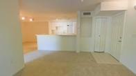 This single-floor space features an open-concept layout that seamlessly connects the living area and kitchen, creating a functional and cohesive environment. The kitchen is outfitted with white cabinetry, a full-size refrigerator, and an oven range, all complemented by a raised breakfast bar that provides a natural visual divider between the cooking and living zones. Track lighting above the kitchen area ensures ample illumination for food preparation and daily tasks. The living area is finished with wall-to-wall carpeting in a warm neutral tone, lending a comfortable, understated character to the space. Tile flooring transitions the entryway and areas adjacent to the kitchen, offering a practical surface near high-traffic zones. A structural column with a built-in recessed niche adds an architectural detail to the open floor plan. A louvered closet door provides convenient storage access. The walls and ceilings are finished in a clean white palette, contributing to a bright and airy atmosphere throughout. Central air conditioning is provided via a ceiling-mounted vent, ensuring year-round climate comfort. Overall, the space presents a clean, neutral canvas well-suited for a variety of uses and furnishing styles.