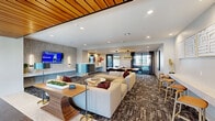 Located at 1345 14th St in Plano, TX, this thoughtfully designed amenity floor spans a generous footprint and offers a comprehensive suite of communal spaces. The open-concept living room serves as the centerpiece, featuring a warm wood-slat ceiling accent, a large wall-mounted TV, plush sectional seating, and patterned carpeting that adds texture and depth. A sleek reception counter anchors the space, which flows naturally into the dining area, illuminated by modern pendant lighting and furnished with a substantial wood-topped communal table and contemporary seating. Bold geometric artwork adorns the walls, enhancing the sophisticated aesthetic throughout. A dedicated office space provides a refined setting for meetings or focused work, outfitted with a marble-topped conference table, upholstered chairs, and large windows offering views of the surrounding urban landscape. A cozy lounge nook with vibrant orange chairs and a decorative pendant light offers an inviting spot for casual gatherings. The fitness center is divided into two well-equipped exercise rooms: one focused on cardio, lined with treadmills, ellipticals, and floor-to-ceiling windows that flood the space with natural light, and a second dedicated to strength training, featuring cable machines, turf flooring, ceiling fans, and energizing motivational wall murals. Together, these spaces create a dynamic, design-forward environment built for both productivity and wellness.