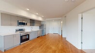 This single-floor unit showcases a clean, contemporary design with a practical and open layout. The kitchen is a standout feature, equipped with a full suite of stainless steel appliances including an electric range, over-the-range microwave, dishwasher, and refrigerator. Sleek gray cabinetry lines the walls, complemented by white countertops and recessed lighting overhead, creating a bright and functional cooking space. The kitchen flows seamlessly into the open living area, which is finished with warm-toned hardwood-style flooring that extends throughout the main spaces, adding continuity and a sense of openness.

A dedicated living zone benefits from a large window that draws in natural light and offers exterior views, with a wall-mounted PTAC unit providing climate control. The bedroom areas are carpeted for comfort and similarly receive natural light through sliding windows with outdoor views. The neutral white walls and clean lines throughout the unit contribute to a bright, minimalist aesthetic. Additional cabinetry is tucked thoughtfully into transitional spaces, offering practical storage without disrupting the streamlined character of the interior.