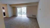 This single-floor residential space offers a clean, functional layout designed for comfortable everyday living. The main living area is generously proportioned, featuring wall-to-wall carpeting in a neutral tone that adds warmth and comfort underfoot. A large sliding glass door serves as a focal point of the room, flooding the space with natural light and providing direct access to a private balcony. The balcony overlooks a verdant, tree-lined setting, offering a serene and secluded outdoor retreat. Sheer white curtains frame the sliding door, softening the natural light and adding a touch of elegance to the otherwise understated aesthetic. The walls are finished in a soft, neutral palette, creating a calm and versatile backdrop suitable for a variety of interior styles. A built-in floor vent provides climate control, ensuring year-round comfort. The overall ambiance of the space is bright, open, and tranquil, with the lush greenery visible through the glass door reinforcing a strong connection to the natural surroundings. The single-floor layout offers ease of movement and accessibility throughout.