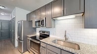 Located at 35 Folly Road Blvd in Charleston, SC, this single-floor unit offers a thoughtfully designed layout with modern finishes throughout. The kitchen serves as a standout feature, showcasing gray shaker-style cabinetry, granite countertops with subtle speckled detailing, and a full suite of stainless steel appliances including a refrigerator, electric range, built-in microwave, and dishwasher. A classic white subway tile backsplash adds a clean, contemporary touch, while track lighting overhead enhances the space's functionality and aesthetic appeal. Light wood-style flooring runs throughout the unit, creating a cohesive and airy feel. The entryway hallway is clean and functional, featuring gray panel doors and ample closet access. The living area benefits from large windows dressed with horizontal blinds, allowing natural light to fill the room, complemented by a ceiling fan with light fixture. The bathroom is well-finished with a tiled tub-and-shower surround in a neutral gray tone, granite-topped vanity cabinetry, and coordinating flooring. The overall color palette — neutral whites, grays, and warm wood tones — creates a modern, cohesive ambiance throughout this Charleston residence.