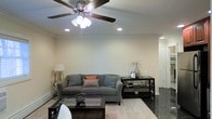 This single-floor space centers around a thoughtfully arranged living room that balances comfort and style. The room is finished with smooth, neutral-toned walls that create a bright and airy feel, complemented by recessed lighting and a ceiling fan with an integrated light fixture, ensuring the space is well-lit and comfortable year-round. Crown molding runs along the upper perimeter of the room, adding a refined architectural detail that elevates the overall aesthetic.

The living area is furnished with a generous gray sofa accented by patterned and solid throw pillows in muted blues and a pop of orange, lending warmth and personality to the space. A glass-top coffee table sits atop a light area rug, grounding the seating arrangement. A dark-finished console table along one wall provides both display and storage space, decorated with vases and framed accents. A floor lamp in the corner adds a soft ambient glow. The flooring transitions from the area rug to what appears to be a dark hardwood or tile near the entryway, offering visual contrast and durability in high-traffic zones.
