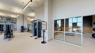 Located at 850 Pacific St in Stamford, Connecticut, this contemporary fitness center is thoughtfully designed to support a comprehensive range of workout routines. The facility is organized across a single level, with distinct zones that cater to both cardio and strength training. The main gym floor is generously proportioned, featuring soaring ceilings and large floor-to-ceiling windows that flood the space with abundant natural light and offer open views of the surrounding area. The room accommodates an extensive array of equipment, including treadmills, ellipticals, cable machines, weight racks, benches, and various strength training machines. Warm wood-toned accent beams and sleek linear pendant lighting contribute to an inviting, modern aesthetic throughout. A separate studio room features full-wall mirrors, a large display screen, yoga mats, medicine balls, and TRX suspension equipment, making it well-suited for group classes, yoga, or personal training sessions. An additional dedicated strength area with rubber flooring offers further free weight and machine options, visible through glass partitions that maintain an open, connected feel. The two-tone accent walls — pairing cool gray with bold golden yellow — add a dynamic visual character to the space. Overall, the fitness center presents a polished, well-equipped environment designed to meet diverse training needs.