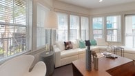 This thoughtfully designed single-floor residence offers a cohesive blend of modern aesthetics and functional living spaces. The open-concept kitchen is a standout feature, showcasing light wood cabinetry, a subway tile backsplash, stainless steel appliances, and a central island with pendant lighting and bar seating — ideal for both cooking and casual dining. Wood-look flooring grounds the space with warmth and continuity.

The living area is bathed in natural light through a sweeping bank of windows fitted with white blinds, creating a bright and airy atmosphere. A neutral color palette with contemporary furnishings lends the space a clean, inviting feel.

The primary bedroom is distinguished by a dramatic floor-to-ceiling upholstered wall panel feature, plush carpeting, and generous windows that invite natural light. A second bedroom offers a serene, minimalist design with vibrant accent curtains and ample natural light.

Both bathrooms are finished with large-format tile flooring, light wood vanities, and white countertops. The primary bathroom features a long dual-sink vanity with an extended mirror, while the secondary bathroom includes a full soaking tub and shower combination. Together, these spaces reflect a consistent commitment to clean lines and modern, comfortable living.