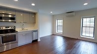 This single-floor space presents a thoughtfully designed open-plan layout that seamlessly integrates the kitchen and living areas. The rich, dark hardwood floors run throughout, adding warmth and character to the interior. The kitchen is outfitted with contemporary flat-front cabinetry in a muted grey tone, complemented by a stainless steel sink and a stainless steel dishwasher, offering a clean, modern aesthetic. Recessed lighting in the kitchen area ensures bright, even illumination for everyday tasks. The living area benefits from natural light streaming through a classic multi-pane window, creating an inviting atmosphere. A wall-mounted ductless air conditioning unit provides efficient climate control. The neutral white walls and white baseboards contribute to a bright, airy feel throughout the space. The overall design balances modern fixtures with classic architectural elements, resulting in a functional and visually cohesive environment suited to a variety of uses.