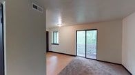 This single-floor space offers a clean, functional layout designed to maximize comfort and usability. The living area is a standout feature, combining two distinct flooring types — smooth hardwood-style planks and plush wall-to-wall carpeting — creating defined zones within the open floor plan. Natural light flows generously into the space through a large sliding glass door that opens directly onto a private balcony, framing lush, green tree views that bring a sense of the outdoors inside. A secondary window further enhances the brightness of the room, ensuring the interior remains well-lit throughout the day. The walls are finished in a neutral tone, providing a versatile backdrop suitable for a variety of interior styles. A textured ceiling adds subtle architectural character to the space. Flush-mounted ceiling lighting provides consistent illumination, complemented by a visible HVAC vent that ensures efficient climate control. Dark baseboards throughout offer a clean contrast against the light-colored walls and flooring. The overall ambiance is calm, airy, and inviting, with a straightforward design that prioritizes livability and a strong connection to the natural surroundings visible through the expansive glazing.