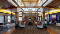 This single-story facility is defined by its warm, lodge-inspired architecture and thoughtfully designed communal spaces. The centerpiece is a grand great room boasting soaring ceilings with exposed structural wood beams and a tongue-and-groove wood ceiling that creates a sense of height and openness. A dramatic floor-to-ceiling stacked-stone fireplace anchors the room, surrounded by a generous seating arrangement of patterned wingback chairs, leather accent chairs, and plush sofas arranged around a central coffee table — ideal for gathering and conversation. Abundant natural light pours in through large windows on multiple sides of the room. Adjacent to the great room is a media lounge area featuring a large wall-mounted flat-screen display and a comfortable sectional sofa with curated artwork on the walls. The entryway welcomes visitors with stone tile flooring, a decorative centerpiece table, arched transom windows above the front doors, and warm wood trim detailing throughout. Wide, well-lit corridors with wood-paneled ceilings and stone tile flooring connect the various areas of the facility, conveying an open and accessible flow. Lush potted greenery is featured throughout, enhancing the inviting and warm ambiance of the entire space.