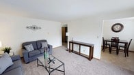 This single-floor space presents a straightforward and functional layout centered around a carpeted living area. The living room features light neutral walls and beige wall-to-wall carpeting that contributes to a warm, understated ambiance. White baseboards frame the perimeter of the room, adding a clean finishing detail. The space is furnished with a grey sofa accented by patterned throw pillows, paired with a glass-top coffee table featuring a black metal frame. A dark wood console table with open lower shelving stands along one wall, complemented by a floor lamp, adding both storage and decorative utility. Decorative metal wall art adds a subtle design element without overwhelming the minimalist aesthetic. A doorway visible toward the rear of the living area leads to an interior hallway, suggesting additional rooms beyond. To the right, a partial view hints at a dining area. The layout feels open and easy to navigate, with ample wall space offering flexibility for furniture arrangement. Overall, the space maintains a clean, move-in-ready appearance with a simple and practical design sensibility suitable for everyday living.
