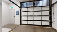 This single-level underground parking facility offers a well-organized and functional space designed to accommodate a variety of vehicles and alternative transportation. Access is provided through a panel-style overhead garage door framed in black steel, featuring translucent upper panels that allow limited natural light into the entry area. A dedicated pedestrian door is located adjacent to the main vehicle entrance, offering convenient access to and from the building interior.

The interior is finished with white-painted concrete walls and structural support columns accented with yellow banding, which also marks the ceiling-level utility runs and delineates traffic flow throughout the space. Parking stalls are clearly defined by yellow line markings on the concrete floor, and the layout accommodates standard passenger vehicles across multiple bays. Overhead fluorescent lighting provides consistent illumination throughout the facility.

A designated bicycle storage area is situated along one wall and includes a multi-bike rack capable of holding numerous bicycles. An additional area appears to accommodate motorcycles and other smaller personal vehicles. Informational signage is posted throughout, including notices indicating that vehicles and contents are left at the owner's own risk. The overall layout is practical and efficiently utilizes the available space.