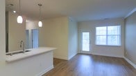 This single-floor space features an open-concept layout that seamlessly connects the kitchen and living areas. The kitchen is equipped with a white countertop island or bar, providing both prep space and a casual seating area. A sleek pendant light hangs above, adding a focused accent to the kitchen zone. Adjacent to the kitchen, a doorway leads to a secondary room or utility space, adding functional versatility to the layout. The living area is finished with warm hardwood-style flooring that runs throughout, lending continuity and a clean aesthetic to the space. Neutral, light beige walls keep the interior feeling airy and open. Natural light is a standout feature, with a large window fitted with blinds and a glass-paneled exterior door that floods the living area with daylight and provides direct outdoor access. Crown molding details along the upper walls add a subtle architectural finish. The overall ambiance is clean, bright, and move-in ready, with a practical open floor plan that maximizes the sense of space.