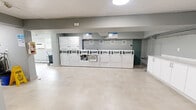 Located at 360 Torrance St in Burlington, Ontario, this shared laundry facility offers a clean, efficiently organized space designed to serve building residents with ease. The single-floor layout is generously proportioned, providing ample room to move comfortably throughout the area.

The main laundry room is the centerpiece of the space, lined with a full row of commercial-grade white washers and dryers arranged along one wall. The light-toned tile flooring runs throughout, contributing to a bright and hygienic atmosphere. Overhead, flush-mount circular lighting ensures the space is well-lit at all times.

A dedicated utility area features white cabinetry with a deep stainless steel farmhouse-style sink, ideal for hand-washing or pre-treating garments. A mounted payment or information panel, wall clock, and television add to the room's functional character. Heating units are wall-mounted, ensuring year-round comfort.

An adjacent windowed section of the facility allows natural light to filter in, with views of surrounding greenery. This area also houses air conditioning units, providing ventilation during warmer months, while baseboard heaters line the walls for cooler seasons.

Access points include multiple doorways with clear signage, and the neutral grey walls throughout lend the space a tidy, modern aesthetic. Overall, this facility is practical, well-equipped, and thoughtfully maintained.