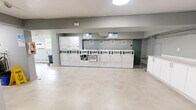 Located at 360 Torrance St in Burlington, Ontario, this shared laundry facility offers a clean, efficiently organized space designed to serve building residents with ease. The single-floor layout is generously proportioned, providing ample room to move comfortably throughout the area.

The main laundry room is the centerpiece of the space, lined with a full row of commercial-grade white washers and dryers arranged along one wall. The light-toned tile flooring runs throughout, contributing to a bright and hygienic atmosphere. Overhead, flush-mount circular lighting ensures the space is well-lit at all times.

A dedicated utility area features white cabinetry with a deep stainless steel farmhouse-style sink, ideal for hand-washing or pre-treating garments. A mounted payment or information panel, wall clock, and television add to the room's functional character. Heating units are wall-mounted, ensuring year-round comfort.

An adjacent windowed section of the facility allows natural light to filter in, with views of surrounding greenery. This area also houses air conditioning units, providing ventilation during warmer months, while baseboard heaters line the walls for cooler seasons.

Access points include multiple doorways with clear signage, and the neutral grey walls throughout lend the space a tidy, modern aesthetic. Overall, this facility is practical, well-equipped, and thoughtfully maintained.