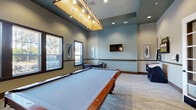 This single-floor game room is thoughtfully designed to accommodate a variety of recreational activities within a spacious, open layout. The room's elevated ceilings create an airy atmosphere, enhanced by a distinctive rectangular pendant light fixture with exposed bulbs that hangs prominently above the central pool table — a full-sized billiard table with a cherry wood frame and light blue felt surface that serves as the room's focal point.

The walls are finished in a two-tone color scheme, featuring soft sage green on the upper portion and a lighter neutral tone below a dark wood chair rail that runs the perimeter of the room. Recessed lighting supplements the pendant fixture, ensuring the space is well-lit throughout. Framed artwork adds subtle decorative touches across the walls.

Natural light enters generously through multiple dark-framed windows fitted with blinds, offering views of surrounding greenery. A wall-mounted flat-screen television is positioned for easy viewing from multiple points in the room.

Additional gaming options include a foosball table and a dedicated seating nook with a dark wood chess and game table surrounded by leather nailhead-trim chairs. The room is finished with a patterned low-pile carpet. A glass-paned French door provides access to the space, completing its polished, amenity-style design.
