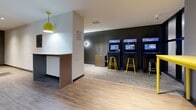 Located at 2840–2850 Carlow Road in Victoria, BC, this dynamic entertainment lounge is a thoughtfully designed recreational space that delivers a lively, social atmosphere for gaming enthusiasts. The single-floor layout is divided into distinct activity zones, each offering its own engaging experience.

The gaming zone is boldly styled with dark grey accent walls, vibrant yellow furnishings, and playful pop-culture wall art — including a iconic "Game On" mural featuring Super Mario and a Pac-Man motif — creating an energetic, arcade-inspired ambiance. Wall-mounted flat-screen TVs and rounded modern light fixtures complement the fun aesthetic, while plush yellow ottomans provide casual seating throughout.

A row of upright arcade cabinets lines one wall, accompanied by yellow bar stools, making it an ideal spot for competitive gaming. The space transitions seamlessly into a billiards area, where a full-size pool table takes center stage beneath a striking industrial pendant light fixture. Adjacent built-in cabinetry, a mounted television, and a wet bar area add functionality and comfort to this zone.

Light wood-look flooring runs through the billiards area, contrasting with the carpeted gaming sections, creating clear visual delineation between zones. A dartboard rounds out the recreational offerings. The overall design balances bold, youthful energy with practical amenities, making this a well-equipped and inviting communal entertainment destination.