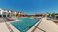 Located at 4271 Woodward Mill Rd in Buford, Georgia, this resort-style outdoor amenity complex is designed to deliver a premium recreational experience. The centerpiece is a generously sized swimming pool with crystal-clear turquoise water, complemented by a separate, shallower wading area equipped with in-water lounge chairs and gentle water features — ideal for relaxing without fully submerging. The pool deck is finished in light-toned pavers that extend across a broad sunning area outfitted with rows of adjustable chaise lounges, providing ample space for residents to unwind under the Georgia sun. A striking wood-framed pergola anchors one end of the deck, offering a shaded retreat alongside the main pool, while a covered stone-and-wood pavilion at the opposite end houses outdoor dining furniture and grilling stations, making it well-suited for social gatherings. Decorative string lighting and landscaped planting beds with young trees add warmth and character to the overall setting. The complex is enclosed by a decorative iron fence, ensuring a sense of privacy, while the multi-story residential buildings visible in the background frame the space with a cohesive community aesthetic. Under open blue skies, the entire outdoor area conveys a bright, welcoming, and upscale atmosphere.