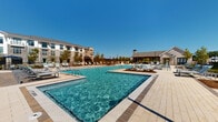 Located at 4271 Woodward Mill Rd in Buford, Georgia, this resort-style outdoor amenity complex is designed to deliver a premium recreational experience. The centerpiece is a generously sized swimming pool with crystal-clear turquoise water, complemented by a separate, shallower wading area equipped with in-water lounge chairs and gentle water features — ideal for relaxing without fully submerging. The pool deck is finished in light-toned pavers that extend across a broad sunning area outfitted with rows of adjustable chaise lounges, providing ample space for residents to unwind under the Georgia sun. A striking wood-framed pergola anchors one end of the deck, offering a shaded retreat alongside the main pool, while a covered stone-and-wood pavilion at the opposite end houses outdoor dining furniture and grilling stations, making it well-suited for social gatherings. Decorative string lighting and landscaped planting beds with young trees add warmth and character to the overall setting. The complex is enclosed by a decorative iron fence, ensuring a sense of privacy, while the multi-story residential buildings visible in the background frame the space with a cohesive community aesthetic. Under open blue skies, the entire outdoor area conveys a bright, welcoming, and upscale atmosphere.