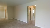 This single-floor space presents a straightforward, functional layout with a clean and neutral aesthetic throughout. The bedroom is finished with wall-to-wall beige carpeting that adds a sense of warmth underfoot, while the white walls and white baseboards create a bright, understated backdrop. The room offers a comfortable sense of space, with enough room to accommodate standard bedroom furnishings.

A doorway leads directly into a walk-in closet, which provides generous storage with visible shelving and hanging space — a practical feature that enhances the room's overall utility. Beyond the closet, an additional room is partially visible, suggesting a connected layout that could serve as a bathroom or secondary space. A window in that adjoining area allows natural light to filter through, contributing to the overall brightness of the interior.

The ceiling is smooth and finished in white, complementing the simple, clean lines of the space. Overhead lighting is present in the closet area, ensuring visibility throughout. Overall, the space offers a well-maintained, move-in-ready environment with a neutral palette that suits a wide range of interior styles.