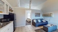 This single-floor unit offers a well-organized layout with distinct living, sleeping, dining, and cooking areas, all united by a charming vaulted ceiling design that lends an airy, open feel throughout. The living area serves as the central hub, furnished with a blue sofa, an upholstered storage ottoman, and a decorative world map accent piece on the wall. A wall-mounted TV and a compact dining table with chairs sit nearby, making the space functional for both relaxing and entertaining. Natural light filters through the windows, brightening the light-toned tile flooring. A wall-mounted HVAC unit provides climate control.

The kitchenette is efficiently arranged along one wall, featuring white cabinetry with open upper shelving, a black refrigerator, a black electric range, and a single-basin sink — offering all the essentials for meal preparation. The bedroom is accessed through a sliding door and features plush carpeted flooring, vaulted ceilings, a queen-sized bed with a upholstered headboard, a nightstand with a lamp, and tasteful wall art, creating a calm and restful retreat. The bathroom is clean and practical, equipped with a tub-shower combination, tile flooring, and chrome fixtures. Overall, the space presents a cohesive, casual aesthetic with nautical-inspired accents throughout.