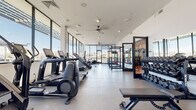 This fitness center is a thoughtfully designed, single-floor facility that combines contemporary architecture with a comprehensive range of workout equipment. The space is organized into distinct training zones, each tailored to a specific type of exercise. Large, floor-to-ceiling curtain-wall windows line the perimeter, flooding the interior with natural light and offering open views of the surrounding area, creating an energizing atmosphere throughout.

The cardio zone features a row of treadmills, elliptical machines, stair climbers, and a Jacob's Ladder-style stair unit, all positioned to take full advantage of the panoramic window views. Overhead ceiling fans contribute to airflow and comfort across the space. The flooring transitions between large-format light grey tiles in the cardio and strength areas, reinforcing the clean, modern aesthetic.

The strength training area is equipped with a full rack of hex dumbbells, a cable machine, a weight bench, and Rogue power racks loaded with bumper plates and medicine balls, accommodating both traditional and functional lifting.

A separate, enclosed studio room features warm hardwood-style flooring, wall-to-wall mirrors, TRX suspension trainers, wall-mounted storage racks with medicine balls and resistance tools, and recessed lighting—ideal for group classes or individual functional training sessions.