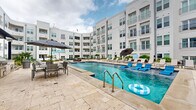 This outdoor amenity space is designed around a generously sized swimming pool that serves as the centerpiece of a vibrant, resort-style courtyard. The pool features classic step entries with stainless steel handrails and is surrounded by a broad travertine stone deck that provides ample room for lounging and entertaining. Rows of chaise lounge chairs and large market umbrellas are positioned along the pool's edges, offering shaded relaxation areas throughout the day.

The courtyard is enclosed on multiple sides by a multi-story building with a crisp white stucco exterior, arched architectural details, and private balconies overlooking the amenity space below. The interior-facing façade creates a sense of privacy and intimacy while enhancing the resort atmosphere.

Beyond the pool, the outdoor space extends into a secondary courtyard area finished in the same travertine pavers. This zone features comfortable outdoor sectional seating upholstered in teal, paired with wicker coffee tables, creating an inviting lounge environment. Additional dining-height table and chair sets are available for al fresco meals. A covered outdoor kitchen area with grilling stations provides a dedicated space for outdoor cooking and entertaining. Tropical plantings, including palm trees and lush greenery, are incorporated throughout, adding color and a vibrant, warm-climate ambiance to the overall setting.