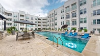 This outdoor amenity space is designed around a generously sized swimming pool that serves as the centerpiece of a vibrant, resort-style courtyard. The pool features classic step entries with stainless steel handrails and is surrounded by a broad travertine stone deck that provides ample room for lounging and entertaining. Rows of chaise lounge chairs and large market umbrellas are positioned along the pool's edges, offering shaded relaxation areas throughout the day.

The courtyard is enclosed on multiple sides by a multi-story building with a crisp white stucco exterior, arched architectural details, and private balconies overlooking the amenity space below. The interior-facing façade creates a sense of privacy and intimacy while enhancing the resort atmosphere.

Beyond the pool, the outdoor space extends into a secondary courtyard area finished in the same travertine pavers. This zone features comfortable outdoor sectional seating upholstered in teal, paired with wicker coffee tables, creating an inviting lounge environment. Additional dining-height table and chair sets are available for al fresco meals. A covered outdoor kitchen area with grilling stations provides a dedicated space for outdoor cooking and entertaining. Tropical plantings, including palm trees and lush greenery, are incorporated throughout, adding color and a vibrant, warm-climate ambiance to the overall setting.