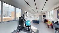 This single-floor space seamlessly combines a fitness area and a casual lounge zone within a clean, modern interior. The fitness section is equipped with a multi-function strength training machine and a stability ball, positioned near expansive windows that frame striking urban skyline views and flood the area with natural light. The lounge portion of the space features a comfortable sofa and casual seating, creating a natural transition from active to relaxed use. A small kitchenette or refreshment station is visible along the far wall, accompanied by a wall-mounted television, adding convenience and functionality. The flooring throughout is a smooth, neutral-toned surface well-suited to both exercise and everyday use. Track lighting runs the length of the ceiling, providing adjustable, even illumination across the entire space. The walls are finished in a crisp white, enhancing the sense of brightness and openness. Large windows along multiple walls ensure consistent natural light and maintain a strong visual connection to the surrounding cityscape. The overall ambiance is clean, contemporary, and practical, designed to support both physical activity and comfortable downtime in an urban setting.
