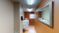 This single-floor shared laundry facility at 3554 Minikahda Ct in St. Louis Park, Minnesota, is designed to serve multiple users with a practical and functional layout. The space features warm wood-style flooring throughout, complemented by a combination of earthy terracotta-orange and neutral taupe wall tones that give the area a distinct, recognizable character.

The laundry area is equipped with front-loading washers and dryers, including Maytag units, and features a utility sink for added convenience. Overhead fluorescent lighting ensures the space is well-lit, and a small window at the far end of the laundry corridor provides a degree of natural light. A large wall-mounted mirror along one side of the corridor enhances the sense of openness in the space.

Adjacent to the laundry area, a corridor leads to a row of individual numbered storage rooms with wood-tone doors, offering residents dedicated secure storage. Windows along this hallway overlook a green outdoor area, adding a connection to the exterior environment.

A separate, more open room with neutral walls and overhead lighting provides additional flexible space, currently furnished with a single chair. The overall layout is straightforward and efficiently organized, prioritizing utility and ease of use for shared occupancy.