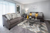 This single-floor residence offers a thoughtfully arranged layout that balances comfort and functionality across its key living spaces. The living room serves as the heart of the home, featuring plush carpeting, a textured ceiling, and a large sliding glass door fitted with vertical blinds that invites natural light and provides access to the outdoors. The room comfortably accommodates a full seating arrangement centered around a coffee table, creating an inviting gathering space. Adjacent to the living room, the galley-style kitchen is outfitted with white cabinetry, light countertops, a coil-top range, and hardwood-style flooring. It opens directly into the dining area, which features a pendant light fixture overhead and a window that brings in additional natural light — ideal for everyday meals. The primary bedroom offers a quiet retreat with carpeted flooring and a textured ceiling, while a secondary bedroom includes a ceiling fan and a bright window, providing flexibility as a guest room or home office. The full bathroom is finished with a wide vanity, ample cabinet storage, hardwood-style flooring, and a shower-tub combination. Neutral gray wall tones and consistent flooring choices tie the space together, creating a cohesive and welcoming atmosphere throughout.
