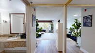 This studio in San Diego, CA (92127) is a smartly designed single-floor space that maximizes functionality within a compact footprint. The open-concept main area seamlessly combines sleeping, living, and kitchen functions, all finished with warm-toned luxury vinyl plank flooring that lends the space a clean, modern feel.

The kitchenette is outfitted with crisp white cabinetry, a single-basin sink, and a countertop microwave, complemented by a full-size refrigerator — providing practical everyday conveniences. A large window with vertical blinds fills the living area with natural light and frames a pleasant green outdoor view, enhancing the sense of openness. A wall-mounted mini-split air conditioning unit ensures year-round climate comfort.

The textured ceiling adds a classic architectural touch, while neutral warm-gray walls create a versatile, calming backdrop throughout. The layout flows naturally from the main living area through a short corridor into the bathroom.

The bathroom is finished with large-format white tile flooring and features a generous granite-topped vanity with an undermount sink, a wide framed mirror with glass shelving, and a private water closet — offering a polished, functional design. Overall, this studio presents a well-organized, efficient layout suited for single-occupancy living in a convenient San Diego location.