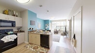 This single-floor unit on Blodgett Drive in Oneonta, New York, offers a well-organized layout that maximizes its compact footprint. The open-concept kitchen and living area form the heart of the space, where light wood-style flooring flows throughout, creating a cohesive and airy feel. The kitchen is equipped with black appliances—including a refrigerator, range, and microwave—complemented by light wood cabinetry, dark granite-style countertops, and a peninsula with a built-in sink and dishwasher. This peninsula serves as a natural divider between the kitchen and the living area, which is brightened by large windows that invite abundant natural light and views of the surrounding area. A bold teal accent wall adds a distinctive visual element to the shared living space.

The bedroom is a cozy, carpeted room featuring a window that provides natural light, offering sufficient space for sleeping and a dedicated study area. The full bathroom features tile flooring, a dark-finish vanity, and neutral wall tones, providing a clean and functional layout. Throughout the unit, the design balances practical living essentials with a modern, welcoming aesthetic suited for everyday comfort.